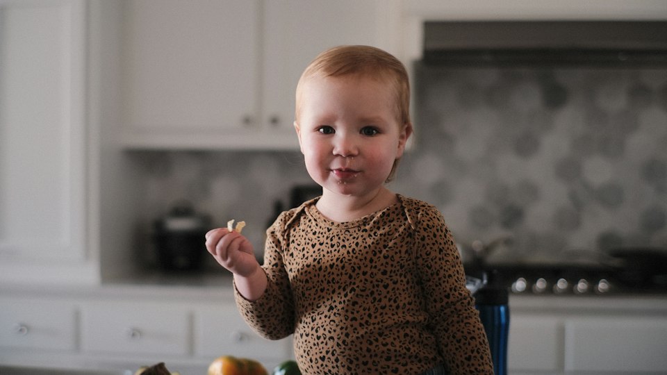 A toddler in a bright home kitchen, holding a piece of food and looking at the camera.