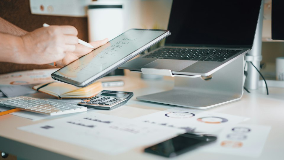 A home desk with a laptop, a calculator and printed charts; someone works through figures on a tablet.